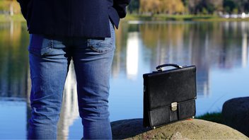 Sicht auf einen See, im Vordergrund ein Mann von hinten, neben ihm seine Aktentasche